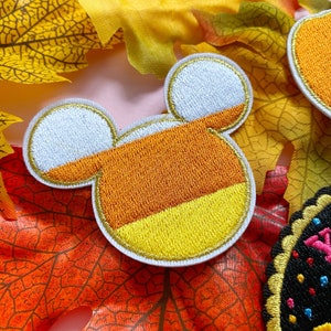 Może przedstawiać: Haftowana naszywka w kształcie uszu Myszki Miki, zaprojektowana tak, aby przypominała cukierki kukurydziane. Naszywka posiada białe, pomarańczowe i żółte sekcje ze złotym wykończeniem. Naszywka jest umieszczona na tle jesiennych liści.