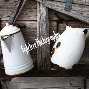 May include: Two white enamel jugs with rust marks hanging on a weathered wooden wall. The jugs are both slightly chipped and have a vintage look.