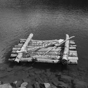 Può includere: Una fotografia in bianco e nero di una zattera di legno fatta di tronchi che galleggia su uno specchio d'acqua calmo. La zattera è fatta di diversi tronchi legati insieme con una corda. I tronchi sono consumati e hanno una consistenza ruvida. L'acqua è calma e riflette il cielo.