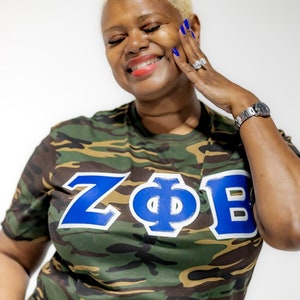 May include: A woman wearing a green, brown, and white camouflage t-shirt with the letters "Z". "Φ", and "B" in blue with white outlines.