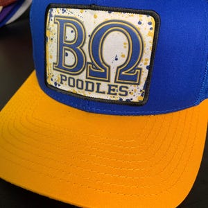 以下が含まれることがあります： 白地に金色の文字で「BQ POODLES」と書かれた白いパッチが付いた青と黄色のベースボールキャップ。背景は白で、金色と青色の斑点模様が描かれている。