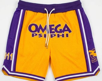 May include: Yellow mesh shorts with purple accents, featuring the text "OMEGA PSI PHI" in purple. The shorts have a drawstring waistband, white and purple trim, and the number "911" on one leg.