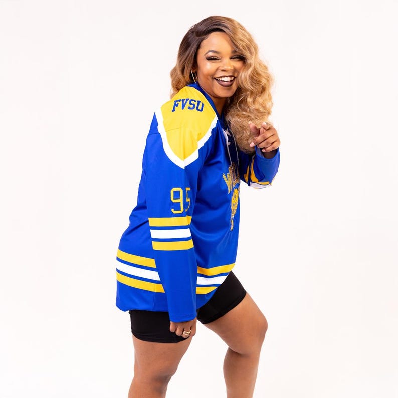May include: A woman wearing a blue and gold hockey jersey with the text "FVSU" and the number "95" on it. She is smiling and pointing to the side.