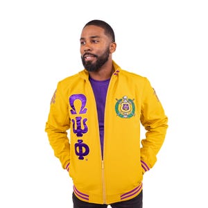 May include: A yellow zip-up jacket with purple and white detailing. The jacket displays the Greek letters Omega, Psi, and Phi in purple on the front. A crest with a shield and crossed swords is embroidered on the chest. The jacket has purple stripes on the cuffs and hem.
