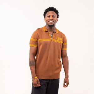 Puede incluir: Polo marrón de manga corta con cremallera y rayas amarillas en el pecho y las mangas. La camisa tiene las letras griegas IΦΘ bordadas en el pecho. La camisa está hecha de punto.