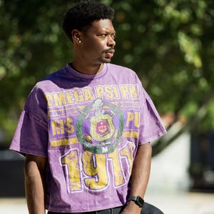 Puede incluir: Una camiseta morada con un acabado desgastado que presenta el texto "OMEGA PSI PHI" y "1911" en amarillo. La camiseta tiene un diseño de escudo en el centro con una espada y un escudo. La camiseta es de manga corta y de corte holgado.