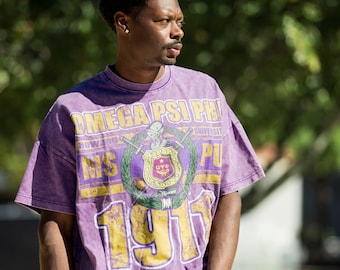 May include: A purple t-shirt with a distressed finish featuring the text "OMEGA PSI PHI" and "1911" in yellow. The shirt has a crest design in the center with a sword and shield. The shirt is short-sleeved and has a relaxed fit.