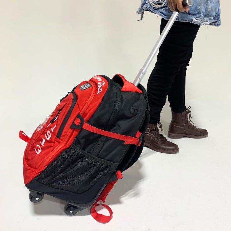 May include: A red and black rolling backpack featuring a retractable silver handle, multiple zippered compartments, side mesh pockets, and black wheels. The front of the bag displays white embroidered text reading 1913 and Zeta Phi Beta.