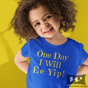 May include: A young child wearing a royal blue t-shirt with the text "One Day I Will Ee-Yip!" in yellow lettering. The child is smiling and looking at the camera.
