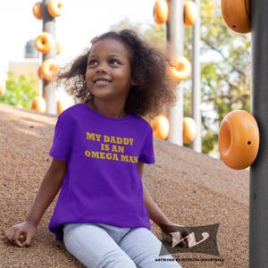 Könnte beinhalten: Ein junges Kind trägt ein lila T-Shirt mit dem Text "My Daddy is an Omega Man" in gelben Buchstaben. Das Kind sitzt auf einer Spielplatzfläche.