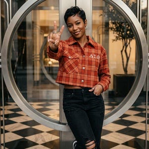 May include: A cropped, orange and red plaid jacket with the Greek letters ΔΣΘ embroidered on the front. The jacket is paired with black ripped jeans. The person is leaning against a glass door with a black and white checkered floor.