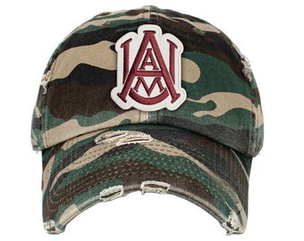 Op de afbeelding: Camouflage baseballpet met een gerafelde rand. De pet heeft een patch met de letters "A" en "M" in bordeauxrood, omlijnd met wit. De hoed is in tinten groen, bruin en beige.