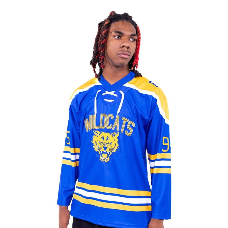 May include: A blue hockey jersey with yellow and white stripes. The jersey has the word "WILDCATS" in yellow letters and a yellow image of a cat with a fierce expression. The numbers "5" and "9" are on the sleeves.