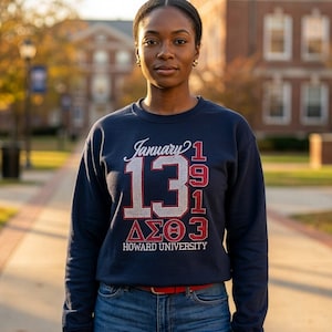 May include: Navy blue sweatshirt with the text "January 13 1919" and "ΔΣΘ" in white and red. The words "HOWARD UNIVERSITY" are printed below. The person is wearing blue jeans and a red belt. The background shows a brick building and trees.