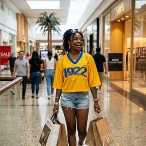 May include: A woman in a yellow jersey with blue trim and the number "1922" is walking in a shopping mall, carrying shopping bags. She is wearing denim shorts, sunglasses, and gold jewelry. The mall has a high ceiling and other shoppers.