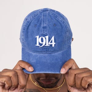 May include: A blue denim baseball cap with the number "1914" embroidered in white. The cap is worn by a person, with the brim partially obscuring their face. The cap has a classic six-panel design and a curved brim.