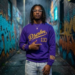 May include: A purple long-sleeve sweatshirt featuring the gold text Psalm 133 on the chest, the number 1911 on the right sleeve, and the text HOW UNIVERSITY on the left sleeve, paired with light blue acid-wash denim jeans and clear glasses.