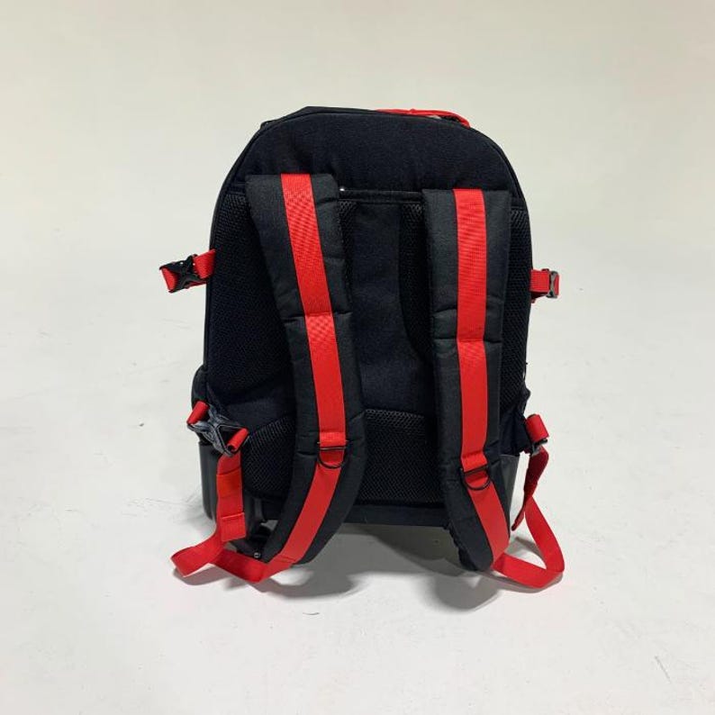 May include: A black backpack featuring bright red adjustable shoulder straps and side compression buckles. The back panel is constructed with black mesh padding, and the design includes integrated red webbing accents along the straps and sides.