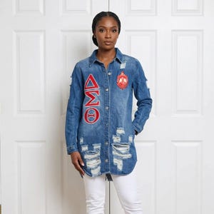 May include: A distressed denim shirt with long sleeves, featuring red embroidered Greek letters and a crest. The shirt has a button-down front and ripped details, paired with white trousers.