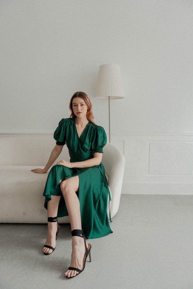 May include: A woman wearing a green wrap dress with puffed sleeves sits on a white couch. She is wearing black heeled sandals with straps.