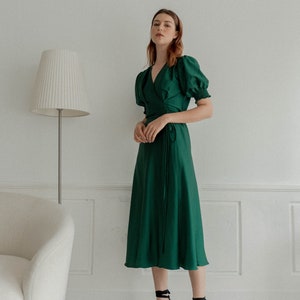May include: A woman wearing a green wrap dress with puffed sleeves and black heeled sandals. She is standing in front of a white wall with a white lamp.