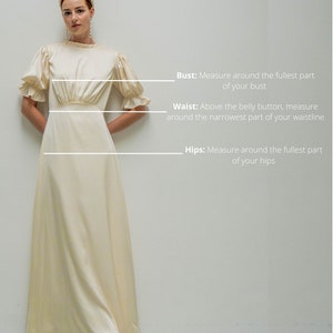 May include: A woman wearing a white satin dress with a ruffled neckline and sleeves. The dress has a fitted bodice and a flowing skirt. The woman is standing in front of a white wall with text describing how to measure bust, waist, and hips.