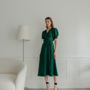 May include: A woman wearing a dark green wrap dress with puffed sleeves and black heeled sandals. The dress has a cinched waist and a flowing skirt.