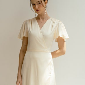 Peut inclure: Une femme portant une robe blanche en satin à encolure en V et manches courtes. Elle porte un bandeau et des boucles d'oreilles en perles.