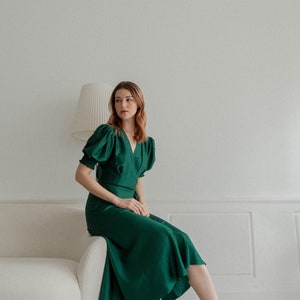 May include: A woman wearing a green satin dress with puffed sleeves and black heeled sandals. She is sitting on a white couch in a white room.