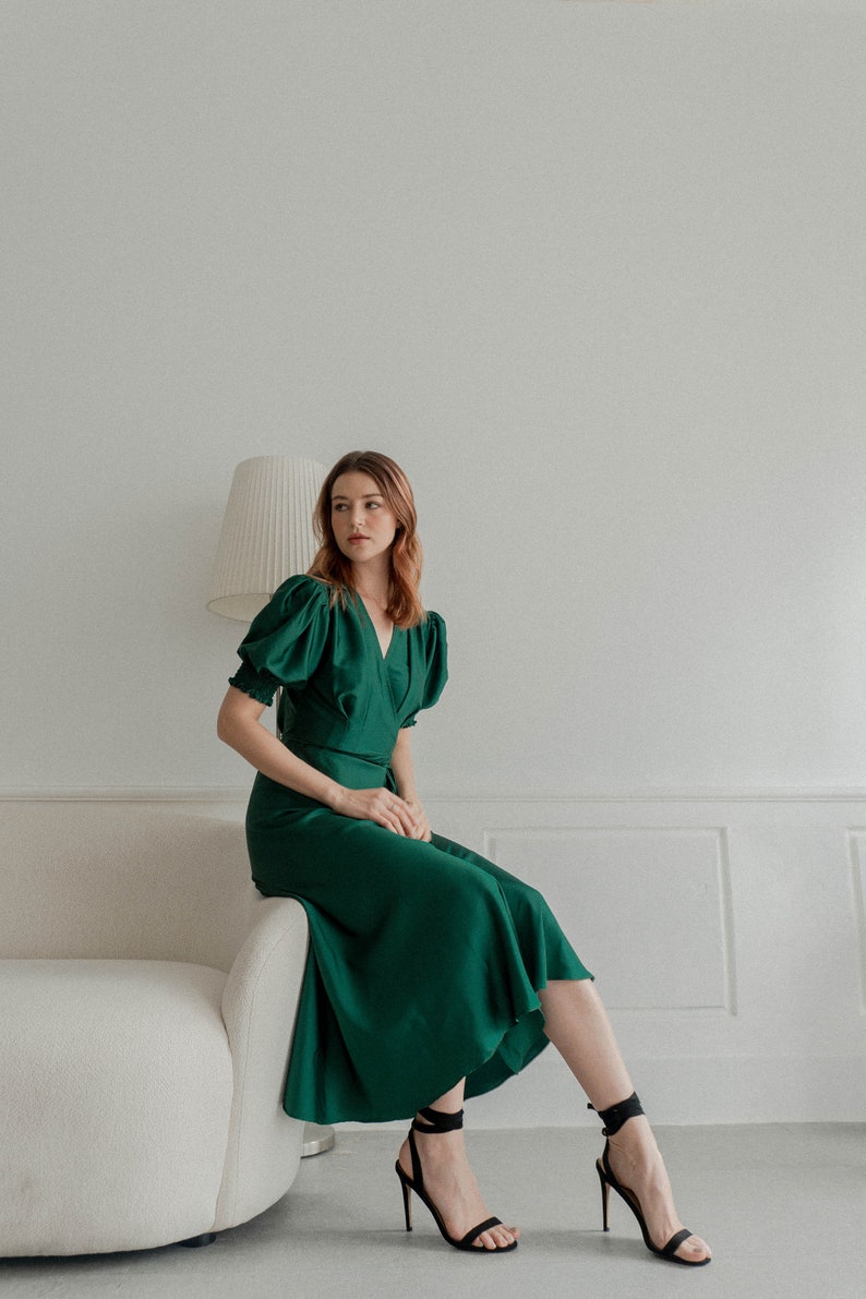 May include: A woman wearing a green satin dress with puffed sleeves and black heeled sandals. She is sitting on a white couch in a white room.