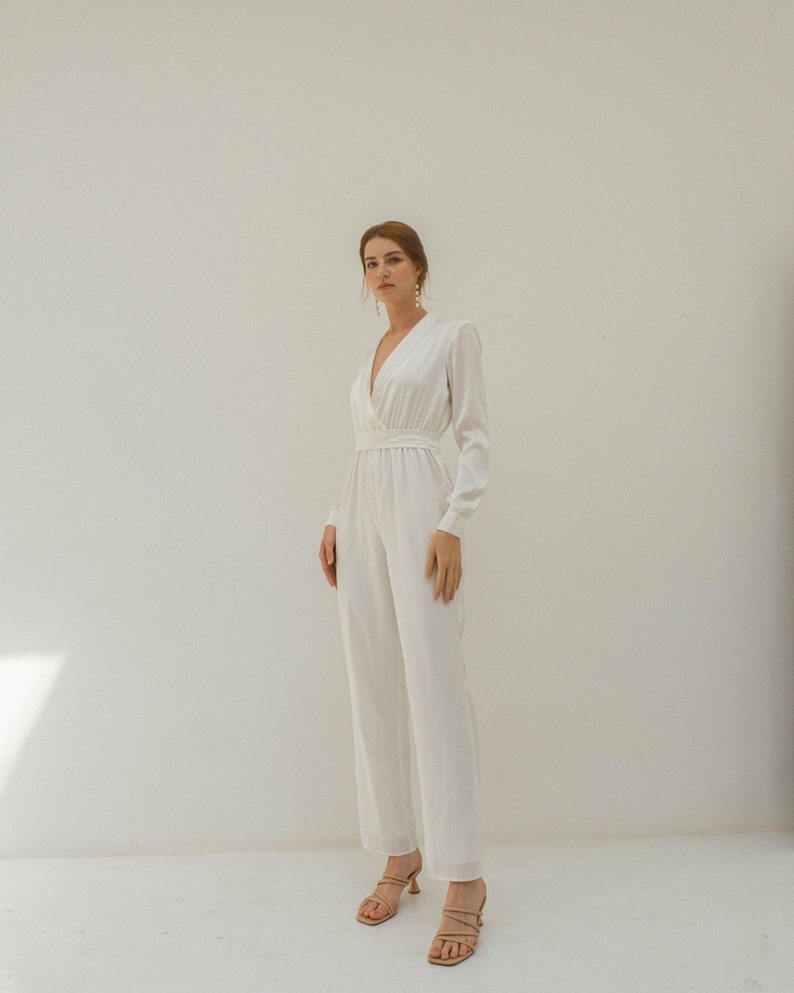 May include: A white long-sleeved jumpsuit with a V-neckline and a fitted waist. The jumpsuit has a relaxed fit and wide legs. The model is wearing the jumpsuit with nude heels.