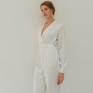 May include: A white satin jumpsuit with a deep V-neckline and long sleeves. The jumpsuit has a fitted bodice and wide-leg pants.