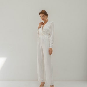 May include: A white long-sleeved jumpsuit with a deep V-neckline and a fitted waist. The jumpsuit has a relaxed fit and wide legs. The model is wearing the jumpsuit with nude heels.