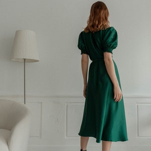 May include: A woman wearing a green midi dress with puffed sleeves and a tie waist. She is wearing black heeled sandals with ankle straps.