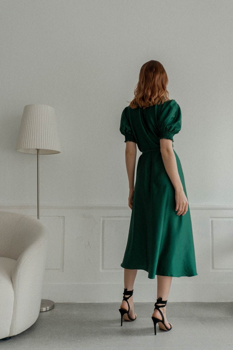 May include: A woman wearing a green midi dress with puffed sleeves and a tie waist. She is wearing black heeled sandals with ankle straps.