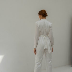 May include: A white satin jumpsuit with long sleeves and a tie at the waist. The jumpsuit has a collared neckline and a relaxed fit.