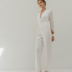 May include: A white long-sleeved jumpsuit with a deep V-neckline and a fitted waist. The jumpsuit has a relaxed fit and wide legs. The model is wearing the jumpsuit with nude heels.
