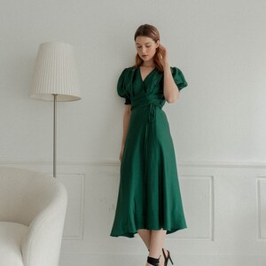May include: A woman wearing a green wrap dress with short puff sleeves and black heeled sandals. She is standing in front of a white wall with a white lamp.
