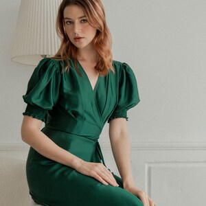 May include: A woman wearing a dark green satin wrap dress with puffed sleeves and a V-neckline. She is sitting on a white couch.