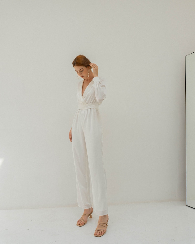 May include: A white jumpsuit with a V-neckline and long sleeves. The jumpsuit has a fitted bodice and wide legs. The model is wearing the jumpsuit with nude heels.