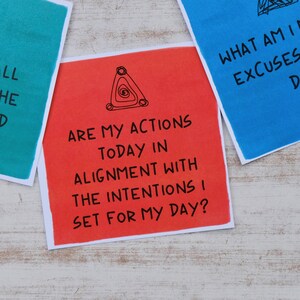 May include: Three colorful cards with a triangle symbol and inspirational questions. The questions are: "What if it all works out the way I wanted it to?", "Are my actions today in alignment with the intentions I set for my day?", and "What am I making excuses not to do?"