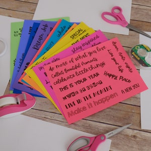 Peut inclure: Une pile de cartes en papier de couleur avec des citations inspirantes &eacute;crites &agrave; l'encre noire. Les cartes sont dispos&eacute;es en &eacute;ventail sur une surface en bois. On voit &eacute;galement des ciseaux, du ruban adh&eacute;sif et d'autres fournitures d'artisanat sur l'image.