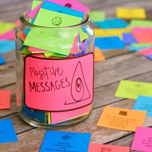 May include: A glass jar filled with colorful sticky notes, labeled "Positive Messages". The jar is surrounded by more sticky notes on a wooden surface.