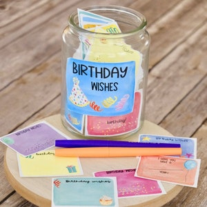 May include: A glass jar filled with colorful cards that say "Birthday Wishes" on them. The jar is sitting on a wooden surface with a few cards scattered around it. The cards have different designs and colors, including pink, blue, yellow, and green. Some of the cards have confetti, stars, and other birthday-themed decorations.