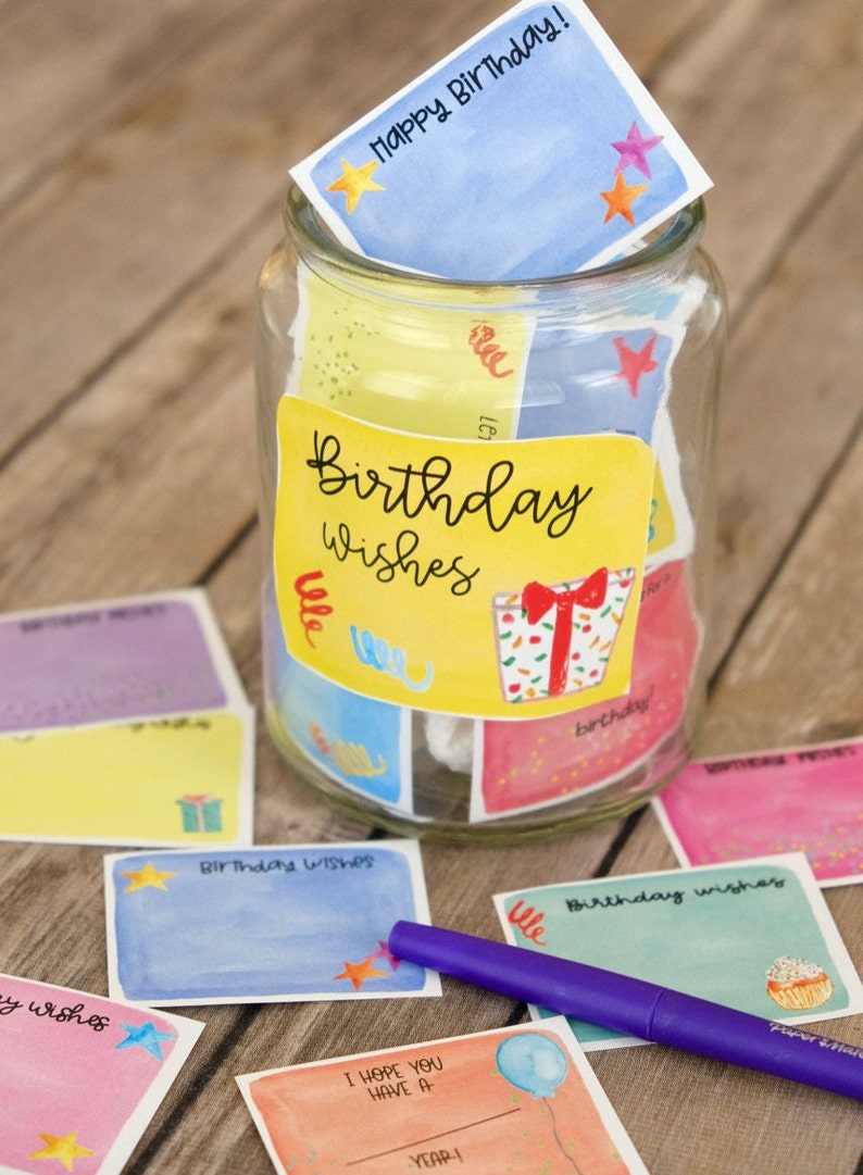 May include: A glass jar filled with colorful cards that say "Birthday Wishes" on them. The jar is decorated with a yellow label that says "Birthday Wishes". There is a purple marker next to the jar.