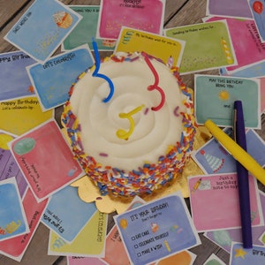 May include: A small white cake with colorful sprinkles and three candles. The cake is surrounded by colorful cards with birthday wishes and messages. The cards have a watercolor style and feature illustrations of balloons, hats, and other birthday-themed imagery.