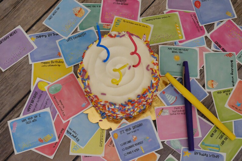 May include: A small white cake with colorful sprinkles and three candles. The cake is surrounded by colorful cards with birthday wishes and messages. The cards have a watercolor style and feature illustrations of balloons, hats, and other birthday-themed imagery.