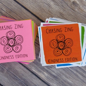 K&ouml;nnte beinhalten: Zwei Stapel bunter Karten mit dem Text "Chasing Zing Kindness Edition" und einem schwarzen und wei&szlig;en Wirbeldesign.