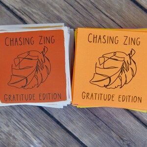 K&ouml;nnte beinhalten: Zwei Stapel orangefarbener und gelber Karten mit dem Text "Chasing Zing Gratitude Edition" und einer schwarzen Strichzeichnung eines Blattes auf jeder Karte.