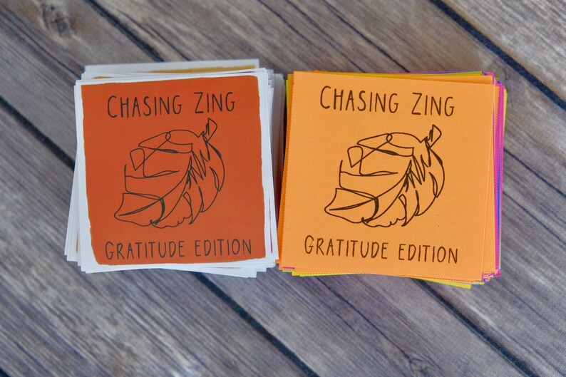 K&ouml;nnte beinhalten: Zwei Stapel orangefarbener und gelber Karten mit dem Text "Chasing Zing Gratitude Edition" und einer schwarzen Strichzeichnung eines Blattes auf jeder Karte.